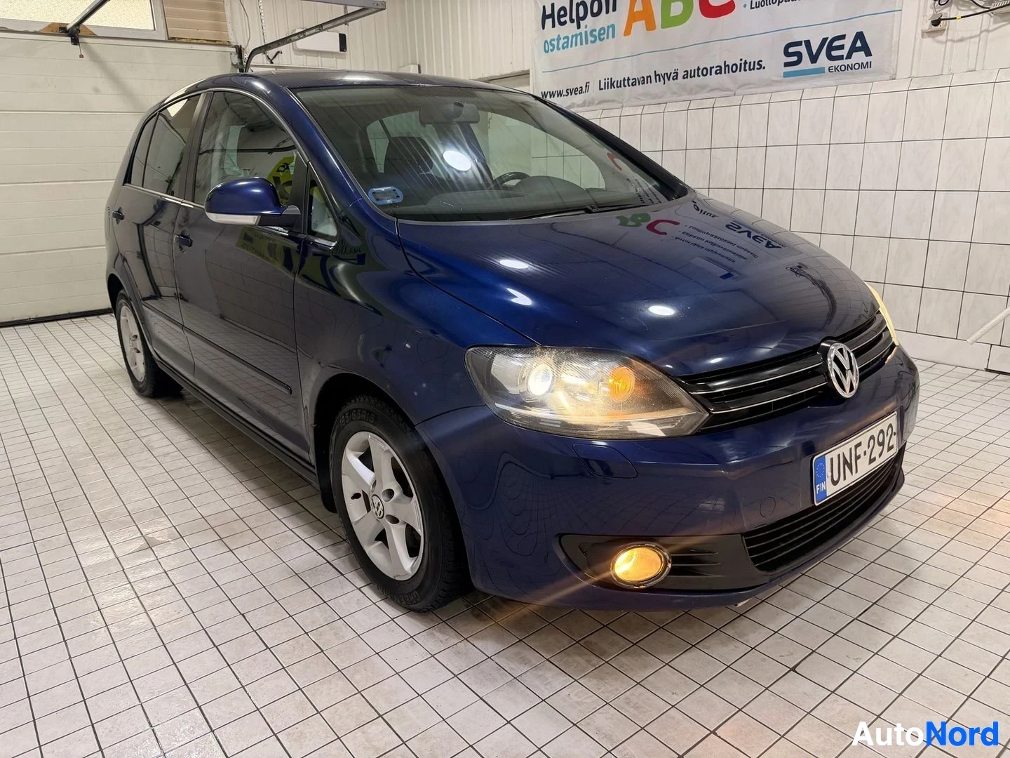 Etuviisto, nokka oikealle: Sininen henkilöauto kuvattuna etuviistosta. Auton etuvalot ja sumuvalot ovat päällä. Auton rekisterikilpi on. Taustalla näkyy mainoskyltti
