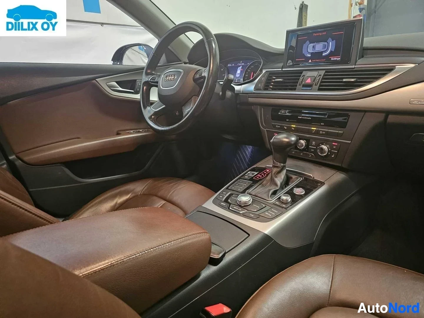 Ohjaamo: Auton sisätila kojelaudan näkökulmasta, ruskea nahkaverhoilu, Audi-ratti, näyttö keskikonsolin yläpuolella, harmaa päätyosa, ilmanvaihto-ohjaimet näkyvissä.