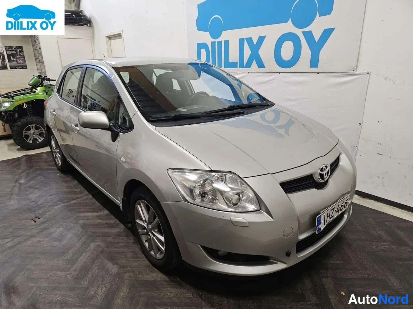 Etuviisto, nokka oikealle: Hopea Toyota Auris -henkilöauto kuvattuna viistosti edestä niin, että keula on kuvan oikeassa laidassa. Toyota-merkki, ajovalot, sumuvalot ja rekisteriki