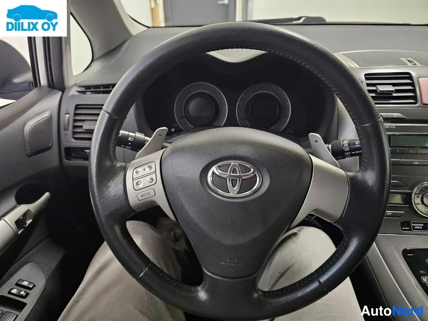Muu: Ohjaamon sisäkuva: Toyota-merkkinen ohjauspyörä vasemmalla ääniohjaimilla ja ratikon äillä, taustalla mittaristo sekä kojelautan radio- ja ilmastointipaneeli.