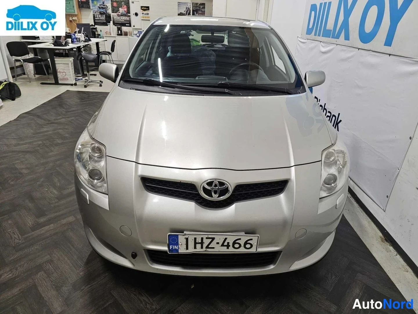 Edestä: Kuvassa on hopeanvärisen Toyotan etuosa. Numeroliitto I HZ-466 ja logo Toyota. Laskeva tuulilasi näkyvissä.