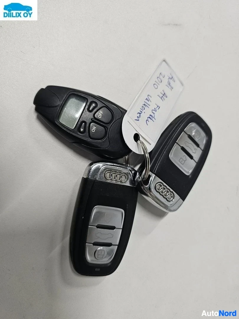 Yksityiskohta: Kolme Audi-merkkistä avaimenperää valkoisella pohjalla. Vasemmalla musta kaukoavain, keskellä hopeanväriset avaimet Audi-logolla, oikealla musta kaukoavain. Val