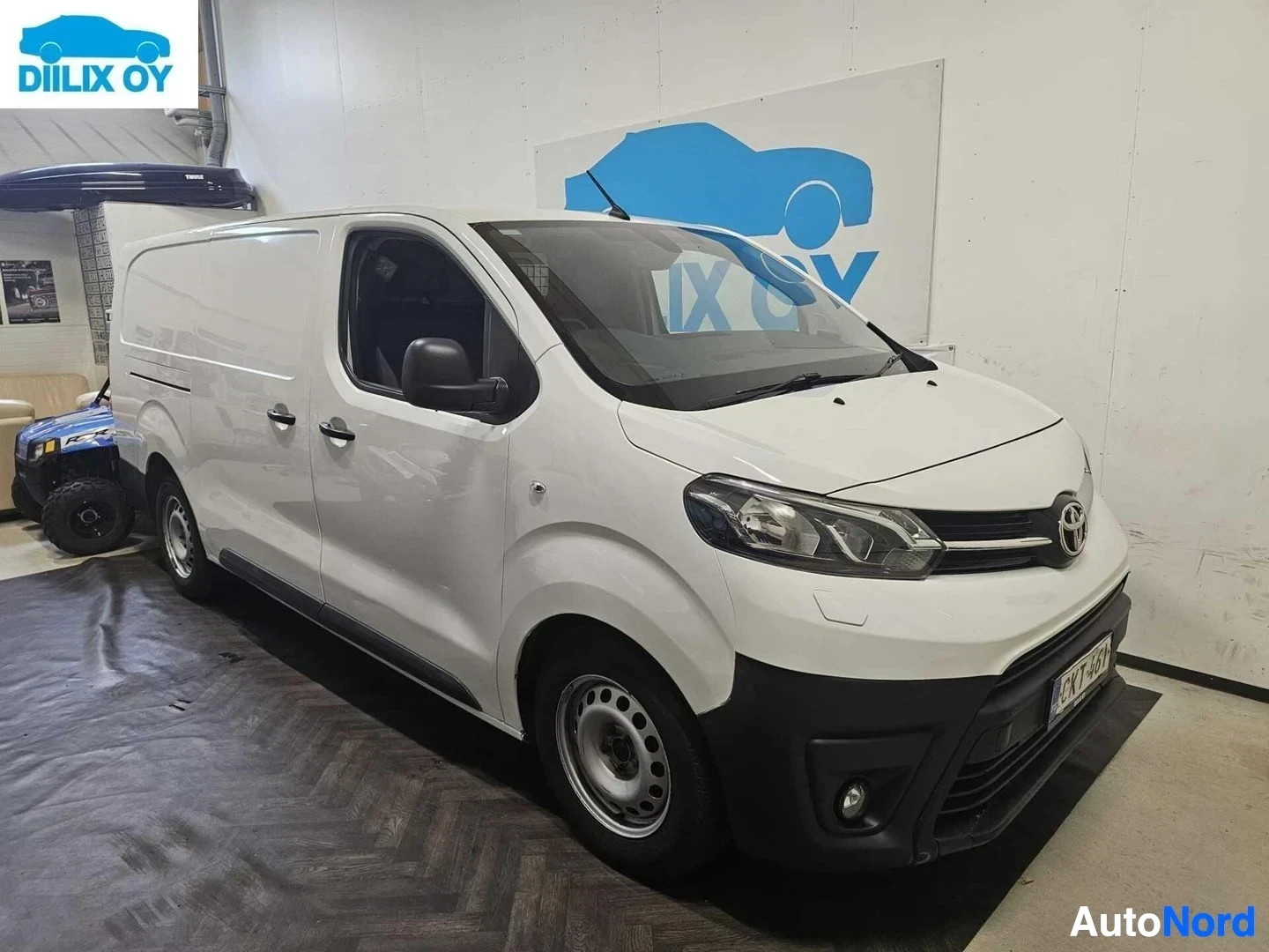 Etuviisto, nokka vasemmalle: Valkoinen Toyota Proace -pakettiauto, jossa näkyy etupuoli ja vasen sivu sisätiloissa vaalealla seinällä.