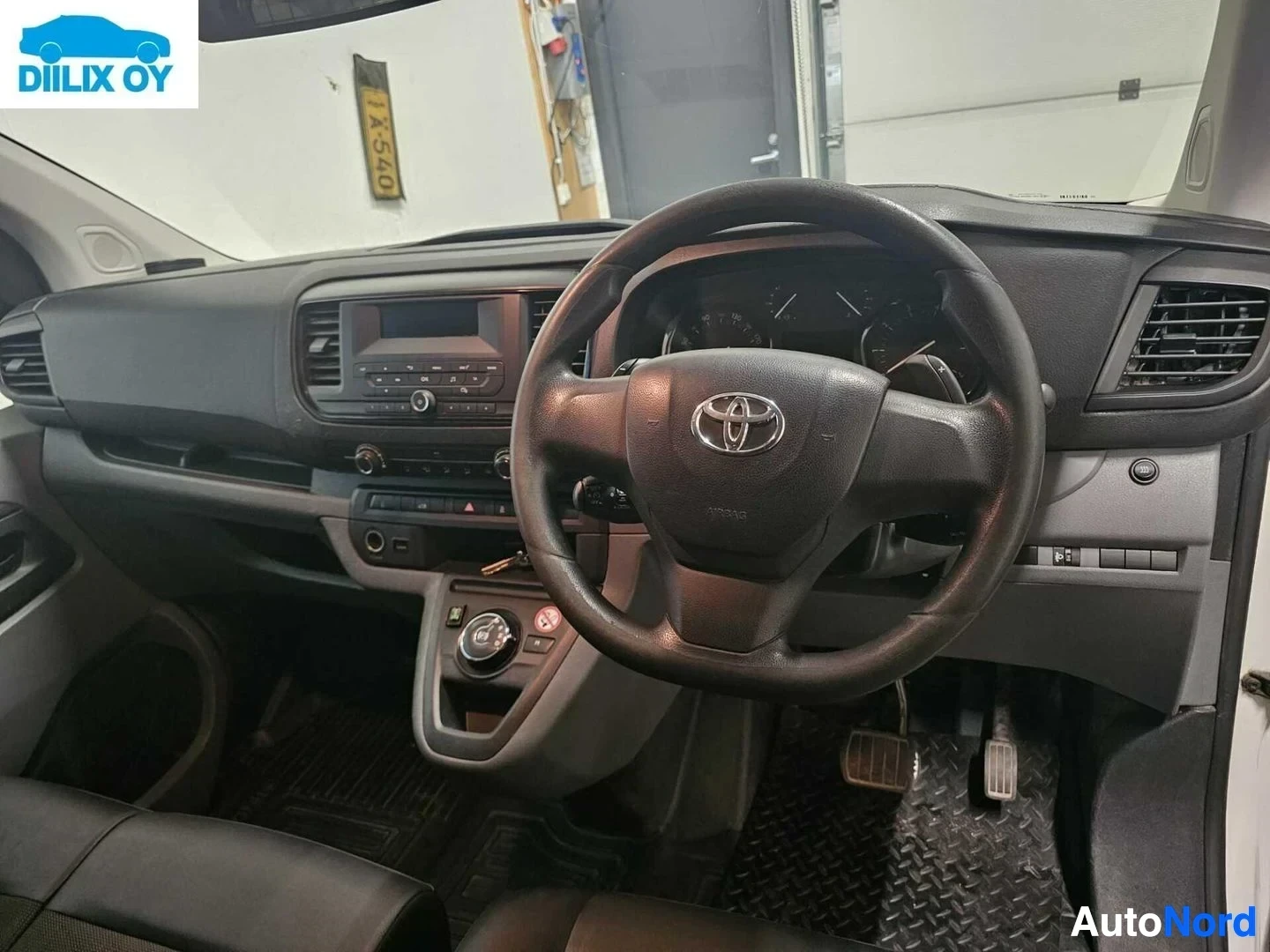 Ohjaamo: Auton ohjaamosta kuvattu kuljettajan näkymä, näkyy tummanruskea ohjauspyörä Toyota-logolla, kojelauta ja säädöt.
