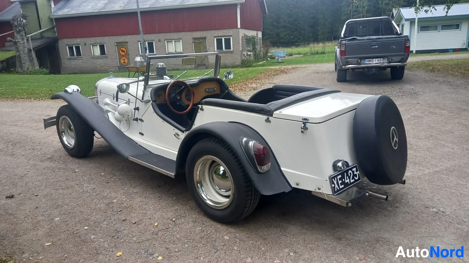 Takaviisto: Kuvan etualalla valkoinen avoauto, mustat lokasuojat ja vararengas perässä. Auton perä on käännetty kuvan vasenta reunaa kohti. Auton rekisterikilpi on XE-423.