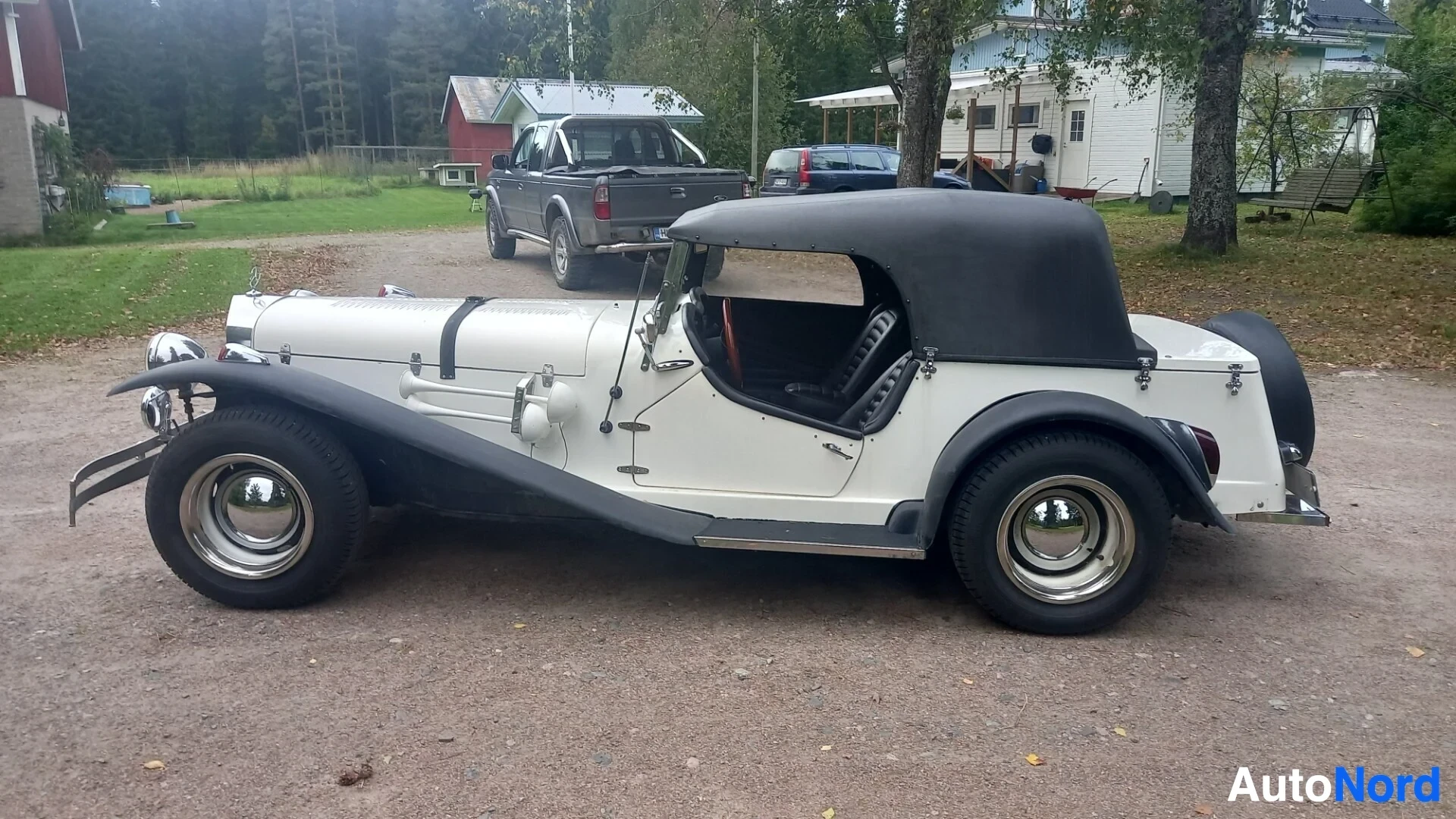 Sivulta: Valkoinen ja musta auto soratiellä, taustalla rakennuksia ja puita.