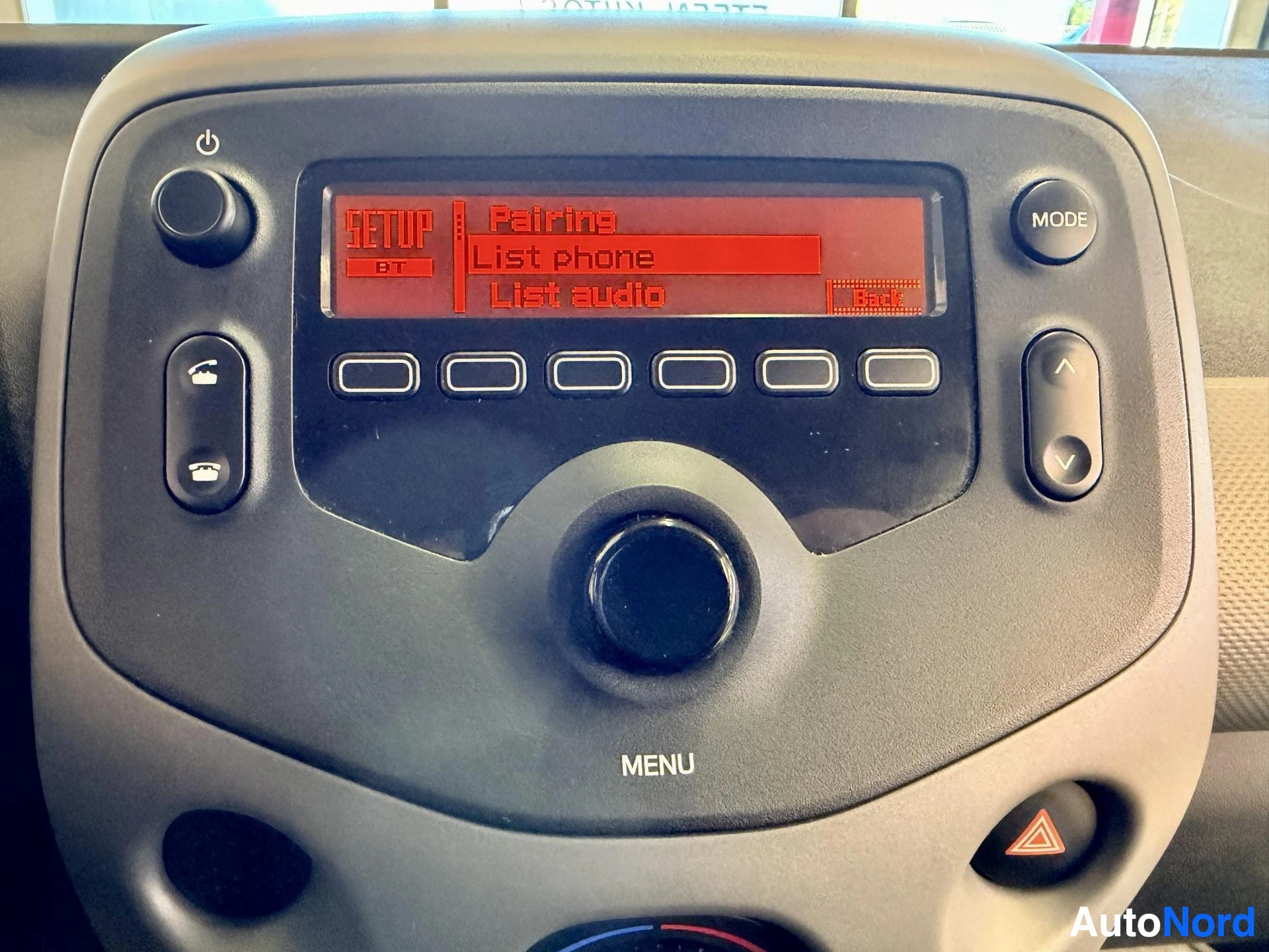 Yksityiskohta: Lähikuva auton keskusnäytöstä, jossa näkyy punaisella tekstillä "SETUP", "Pairing", "List phone" ja "List audio". Useita painikkeita ja kiertosäädin ympäröivät