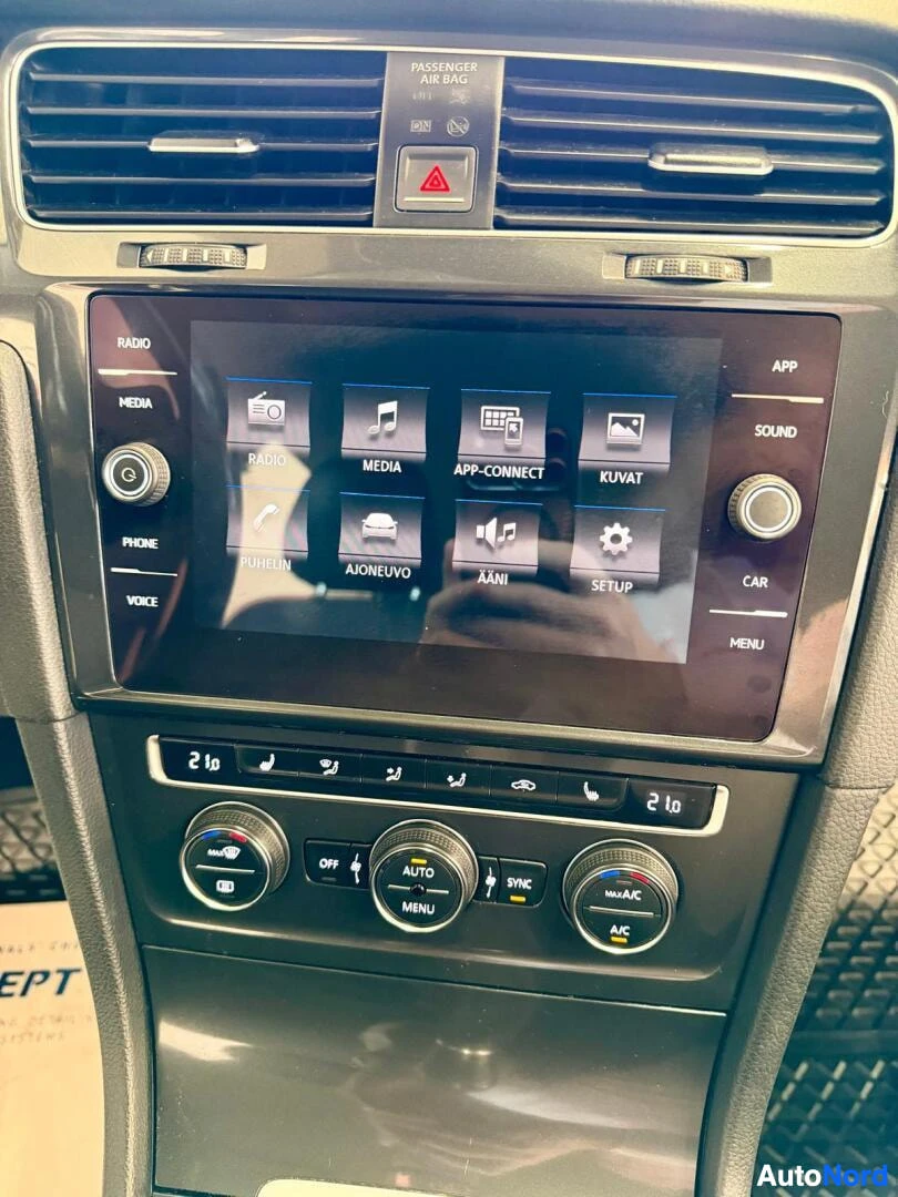 Ohjaamo: Kuva auton sisätiloista, jossa näkyy infotainment-järjestelmä ja ilmastoinnin säätimet. Infotainment-järjestelmä näyttää kuvakkeita, kuten Radio, Media, App-Con