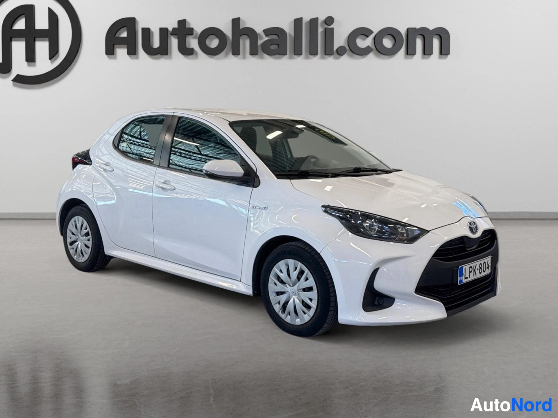 Etuviisto, nokka oikealle: Valkoinen Toyota Yaris edestä ja vasemmalta sivulta kuvattuna. Auton rekisterinumero on