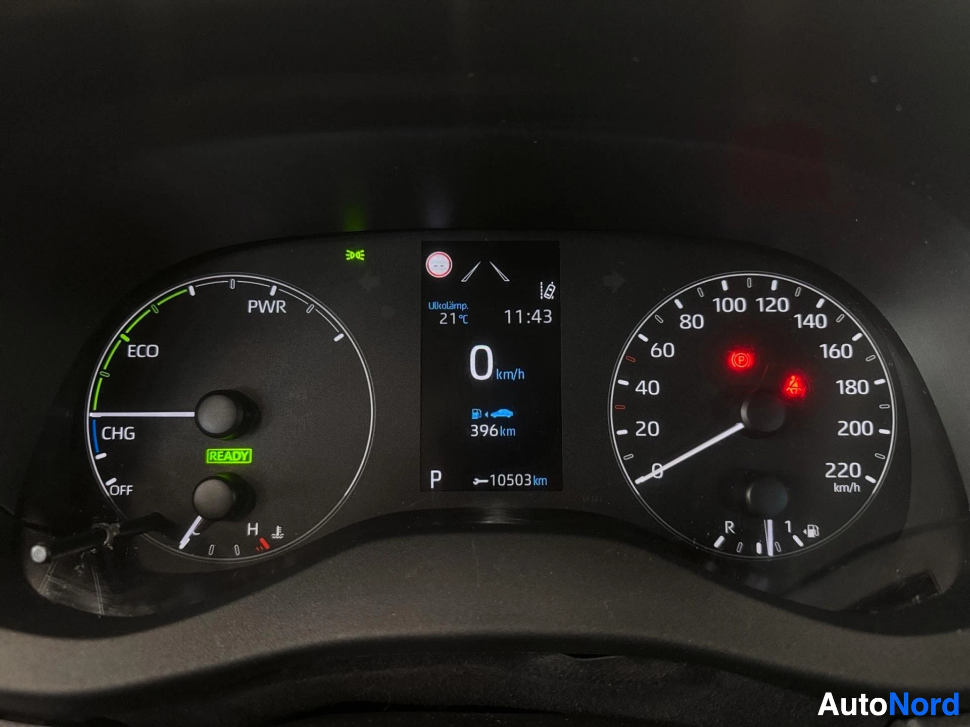 Yksityiskohta: Lähikuva auton mittariston näytöstä, jossa näkyvät nopeusmittari (0-220 km/h), tehomittari (CHG-ECO-PWR), polttoainemittari, ulkolämpötila (21°C), aika (11:43),