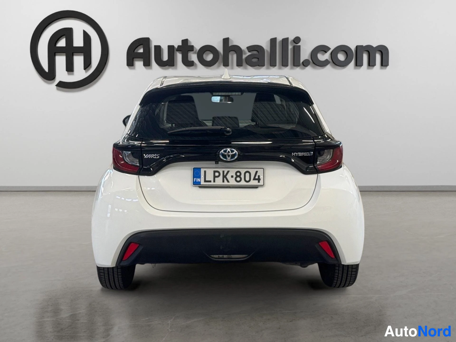 Takaa: Valkoinen Toyota Yaris näkyy suoraan takaa. Auton takarekisterikilpi on. Takaikkunan alareunassa on Toyota-logo ja teksti Hybrid