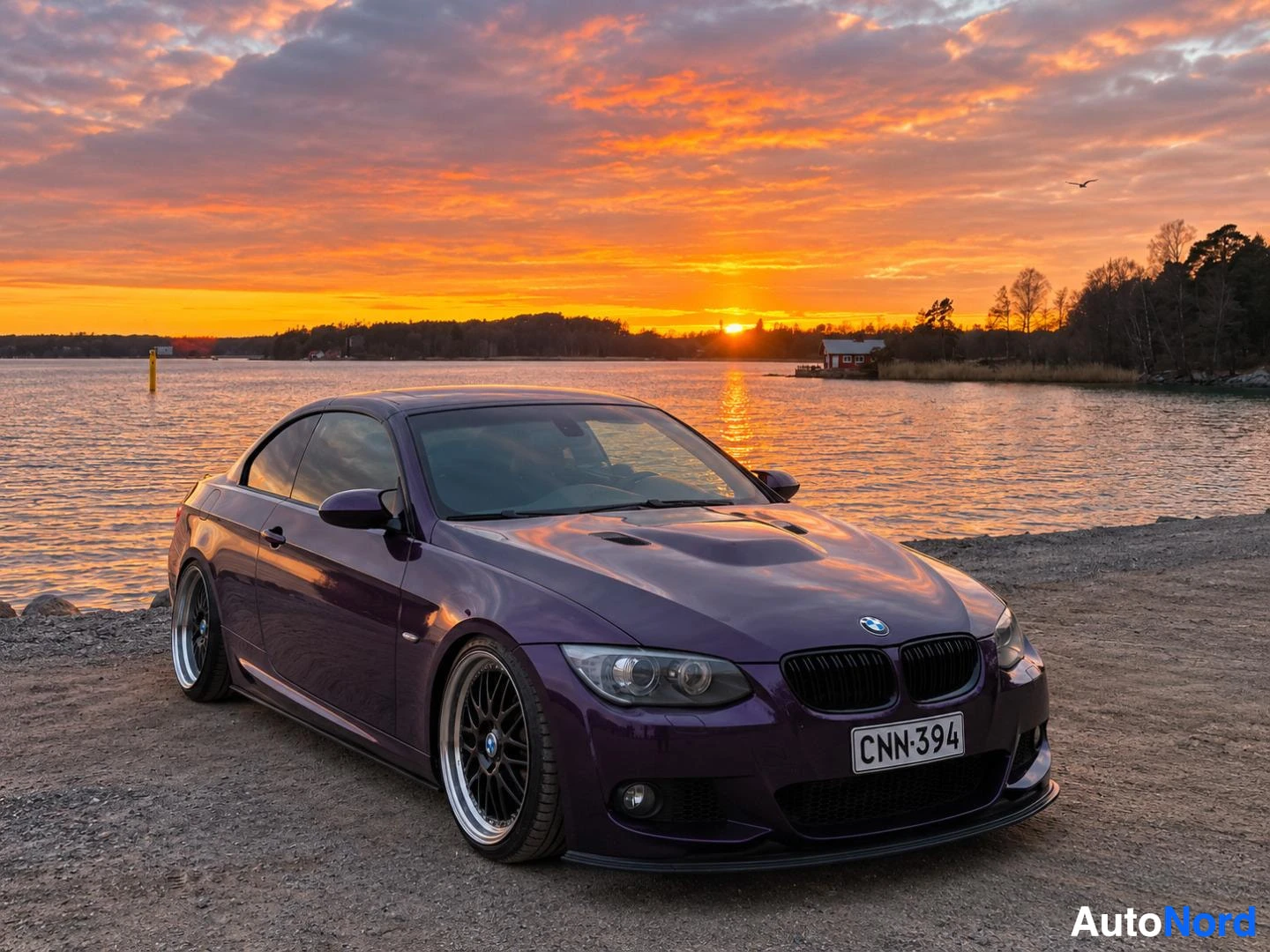 Etuviisto, nokka oikealle: Violetti BMW-henkilöauto, rekisteritunnus, pysäköitynä vesistön rannalle auringonlaskun aikaan. Autossa on mustat monipuolaiset kevytmetallivante