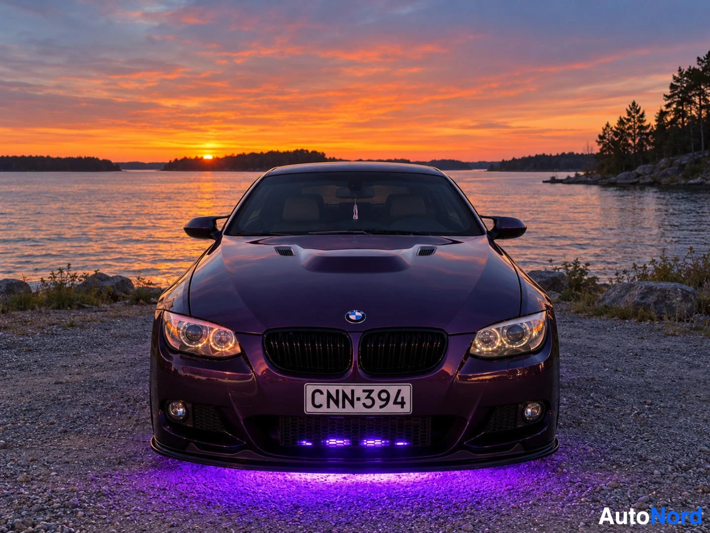 Edestä: Violetti BMW-henkilöauto edestä päin kuvattuna auringonlaskun aikaan vesistön rannalla. Autossa on musta etusäleikkö ja etupuskurin alla violetti valaistus. Rek