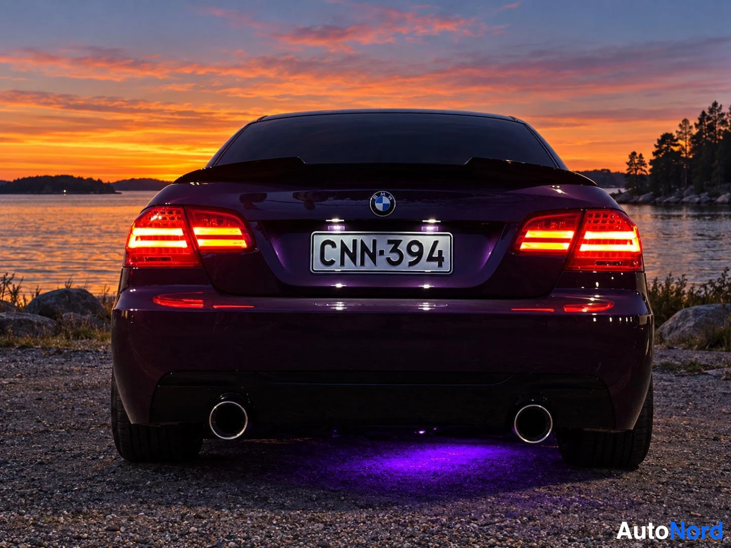 Takaa: Violetti BMW-henkilöauto takaa päin kuvattuna auringonlaskun aikaan vesistön rannalla. Autossa on kaksi pakoputken ulostuloa ja takapuskurin alla violetti valai