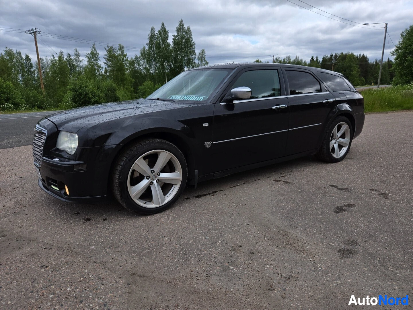 Etuviisto, nokka vasemmalle: Musta Chrysler 300C Touring -farmariauto pysäköitynä tien sivuun, etuvasen kolme neljäsosaa kuvassa, alumiinivanteet edessä ja takana.