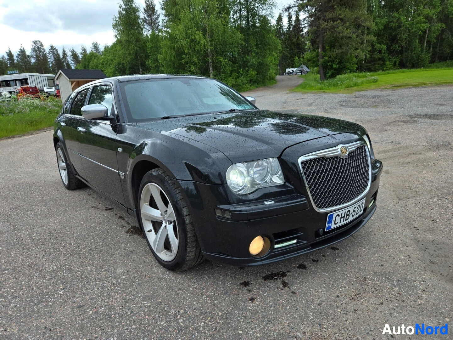 Etuviisto, nokka vasemmalle: Musta Chrysler 300C farmari seisoo asfalttitiellä, etuosa ja oikea kylki näkyvät. Auton edessä näkyy rekisterikilpi