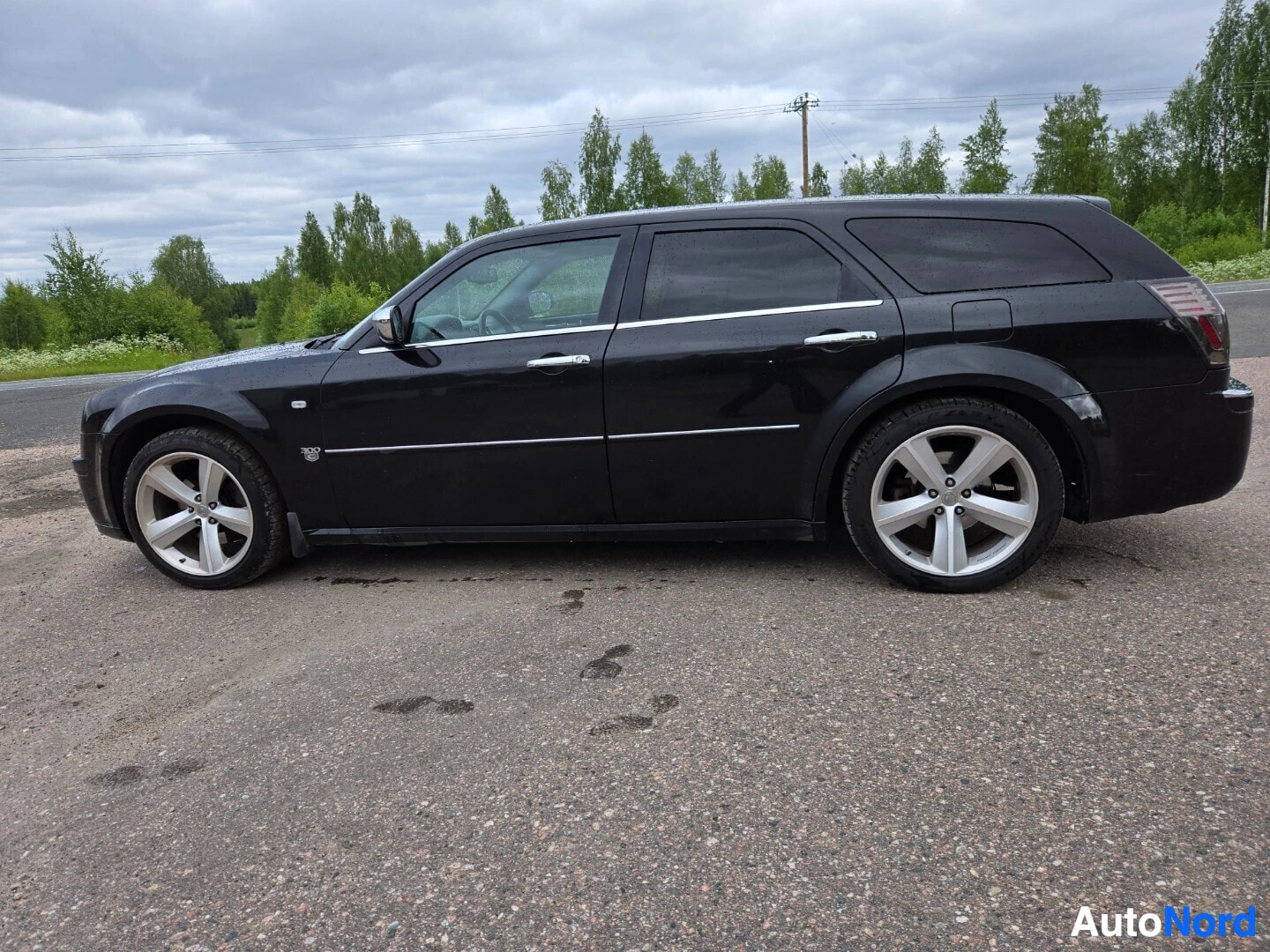 Sivulta: Musta Chrysler 300C Touring -farmariauto pysäköitynä tien sivuun, vasen kylki kuvassa, alumiinivanteet edessä ja takana.