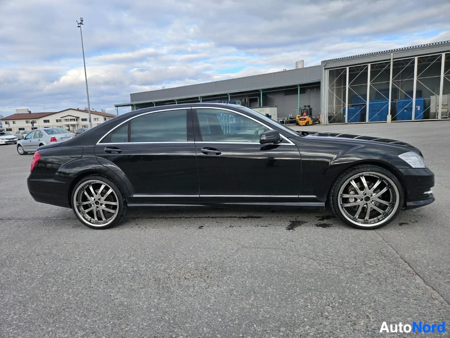 Sivulta: Musta sedan-tyyppinen auto parkkipaikalla, vasen kylki kokonaan näkyvissä. Autossa on kromivanteet.