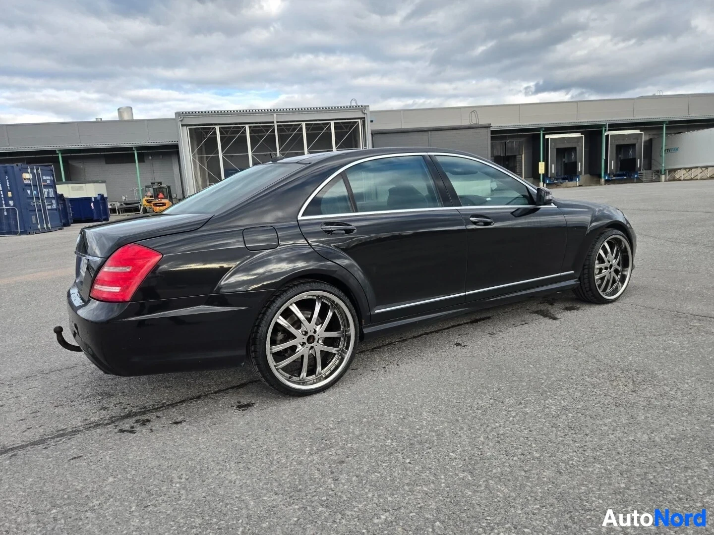 Takaviisto: Musta sedan-tyyppinen auto parkkipaikalla, takanurkasta kuvattu. Vasen kylki ja takapää näkyvissä. Autossa on kromivanteet ja vetokoukku.