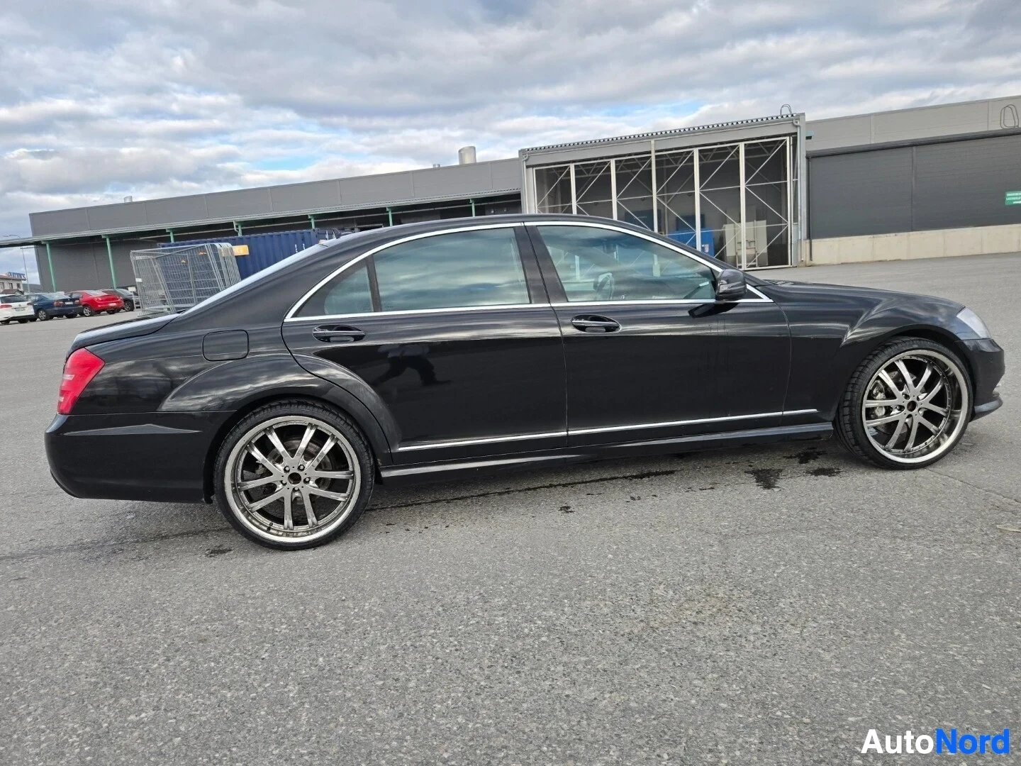 Takaviisto: Musta sedan-tyyppinen auto parkkipaikalla, vasen kylki ja takaosa näkyvissä. Autossa on kromivanteet.