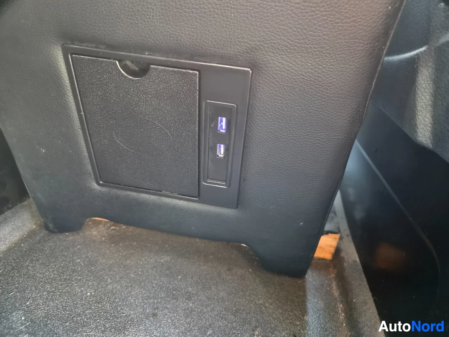Yksityiskohta: Lähikuva takaistuimen selkänojan takaosassa olevasta USB-latausporttipaneelista, jossa on kaksi USB-A-porttia, toisessa sininen valo. Vieressä on musta säilytys
