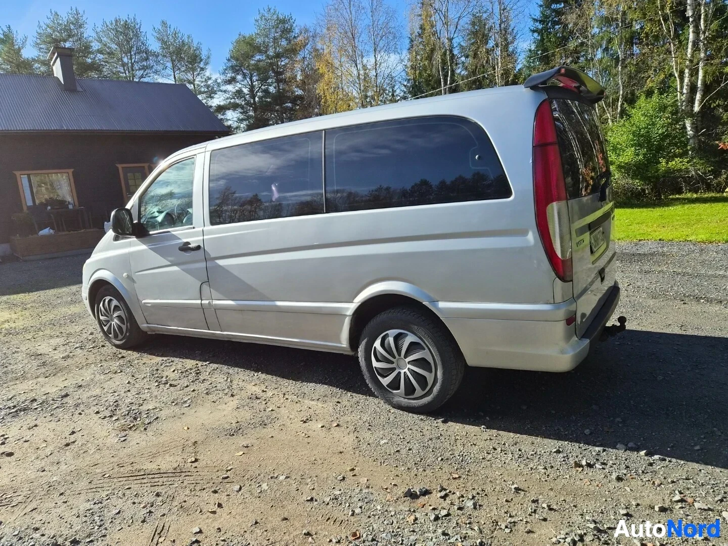 Takaviisto: Hopeanvärinen pakettiauto takaa ja oikealta sivulta kuvattuna hiekkatiellä, jossa rakennus ja metsä taustalla. Autossa on kattospoileri ja vetokoukku. Takana ja