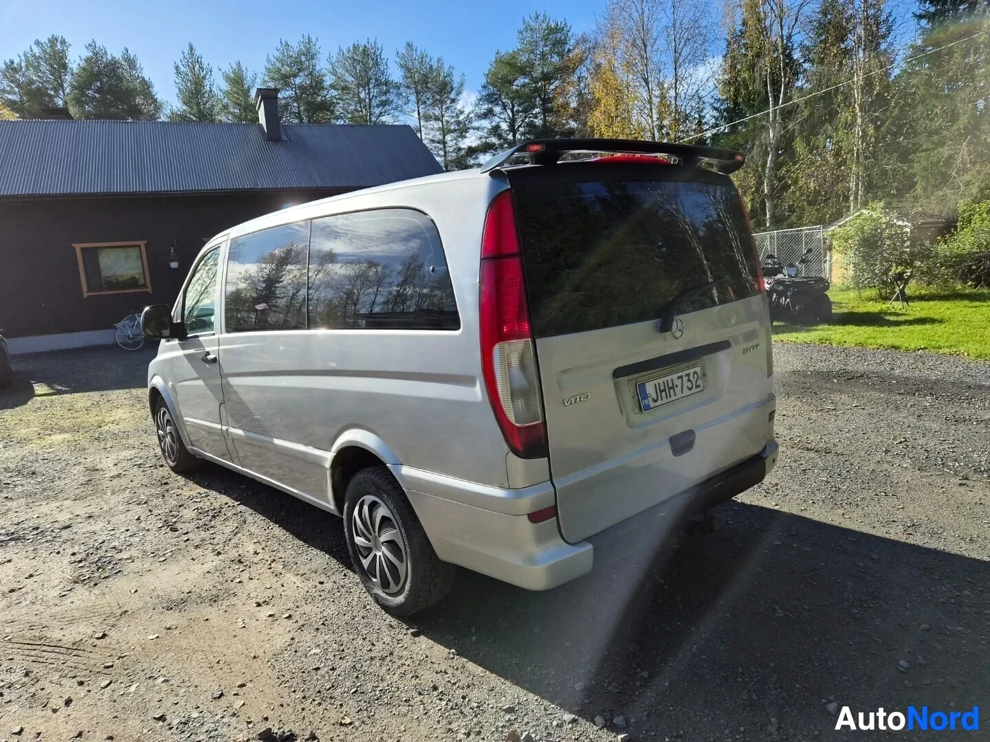 Takaviisto: Hopeanvärinen pakettiauto takaa ja oikealta sivulta kuvattuna hiekkatiellä, jossa rakennus ja metsä taustalla. Autossa on kattospoileri ja vetokoukku. Takana nä