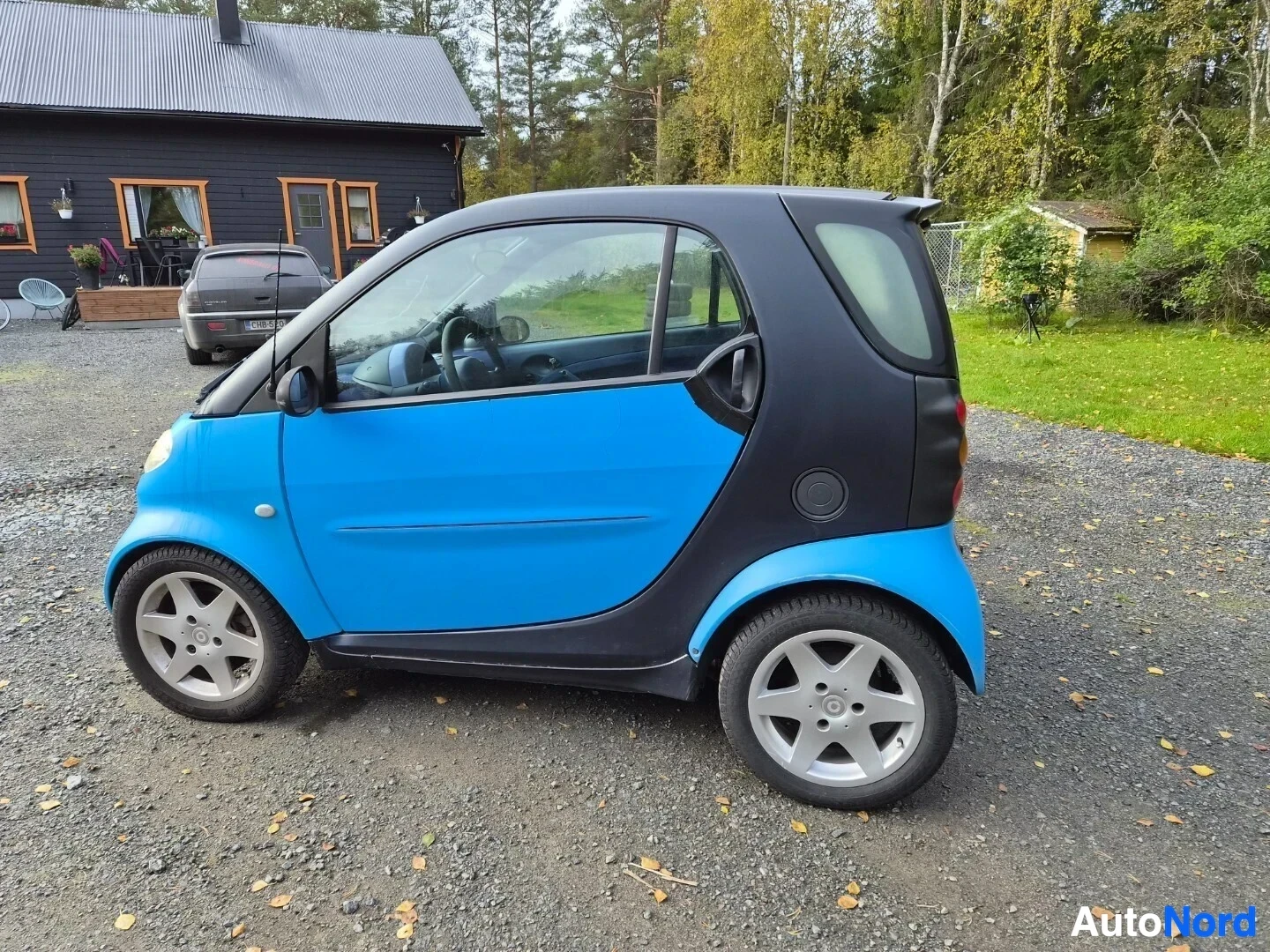 Takaviisto: Siniset ja mustat koripaneelit omaava auto pysäköitynä hiekkapihalle, vasen kylki ja takaosa näkyvissä. Auton alla on harmaat alumiinivanteet.