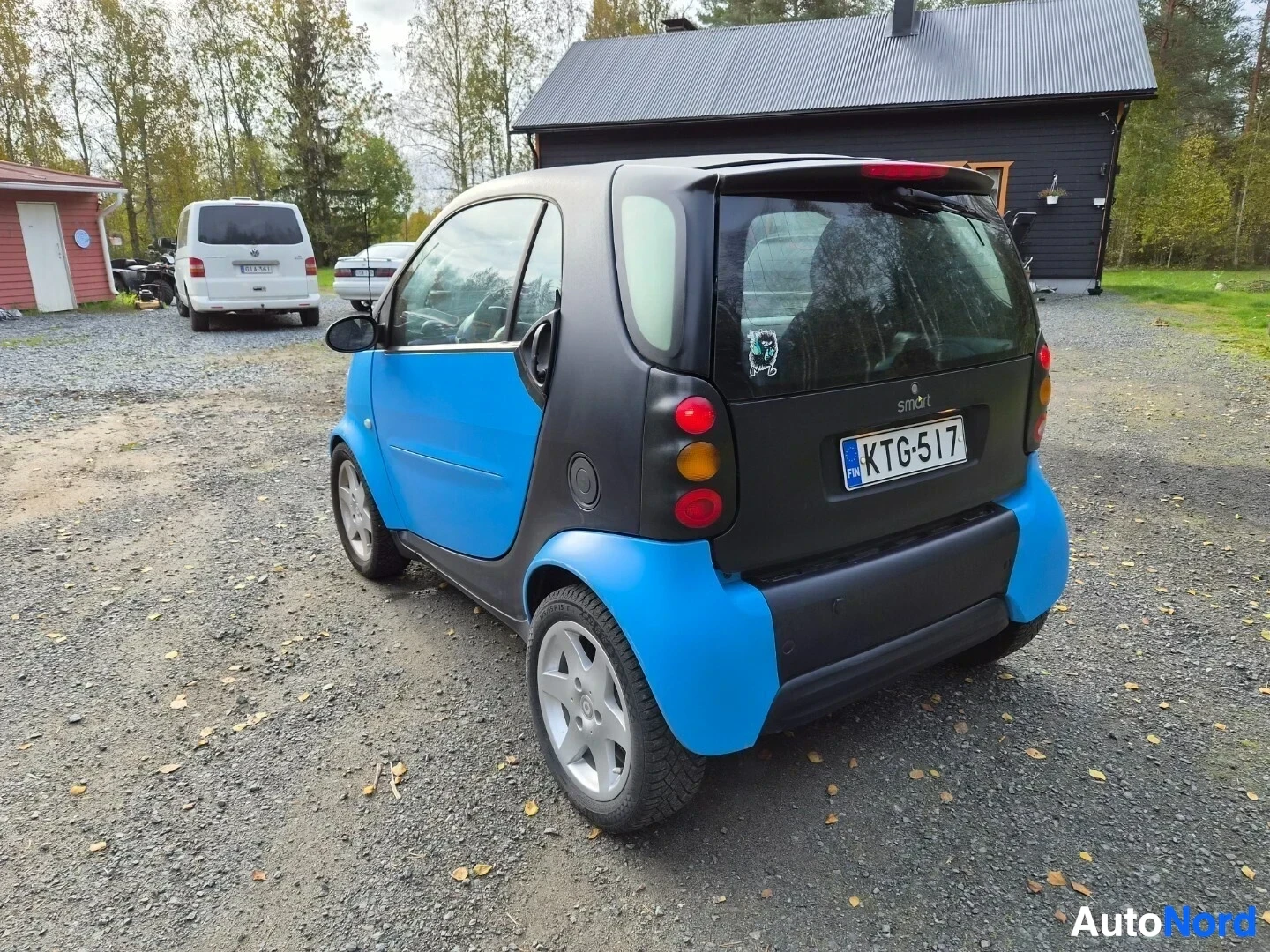 Takaviisto: Smart Fortwo -henkilöauton takavasemmalta otettu kuva, jossa ajoneuvo on pysäköity hiekkakentälle. Autossa on siniset ja mustat paneelit, ja siinä on suomalaine