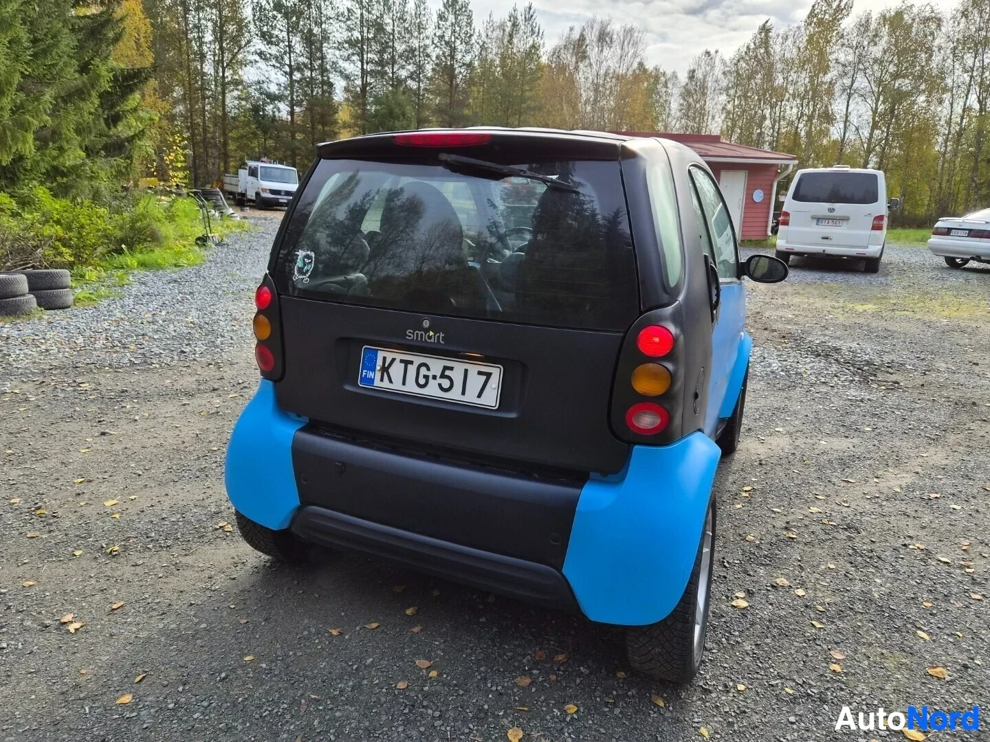 Takaviisto: Smart Fortwo -henkilöauton takaa oikealta otettu kuva, jossa ajoneuvo on pysäköity hiekkakentälle. Autossa on siniset ja mustat paneelit, ja rekisterikilpi KTG-