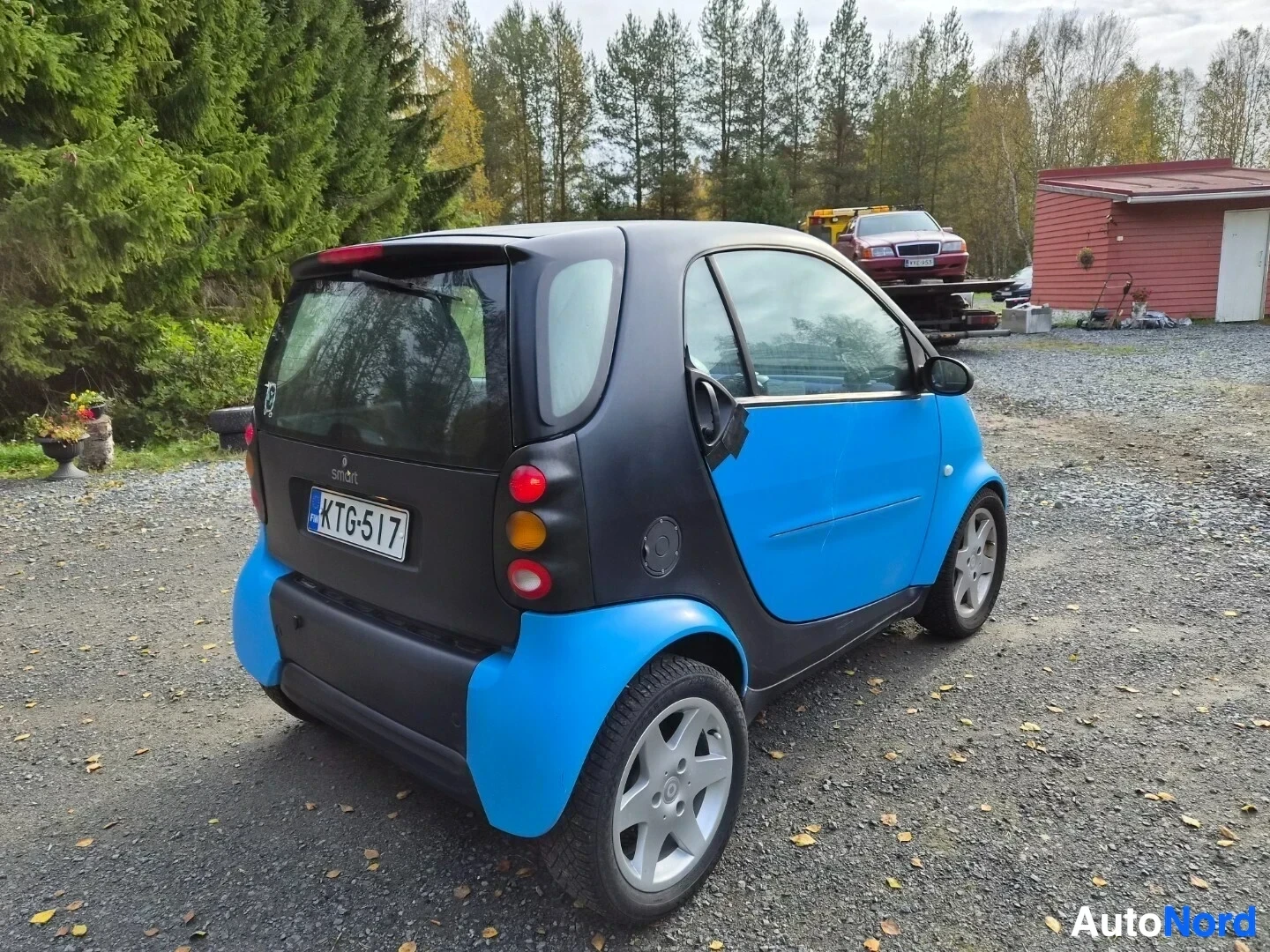 Takaviisto: Smart Fortwo -henkilöauton takavasemmalta otettu kuva, jossa ajoneuvo on pysäköity hiekkakentälle. Autossa on siniset ja mustat paneelit, ja siinä on suomalaine