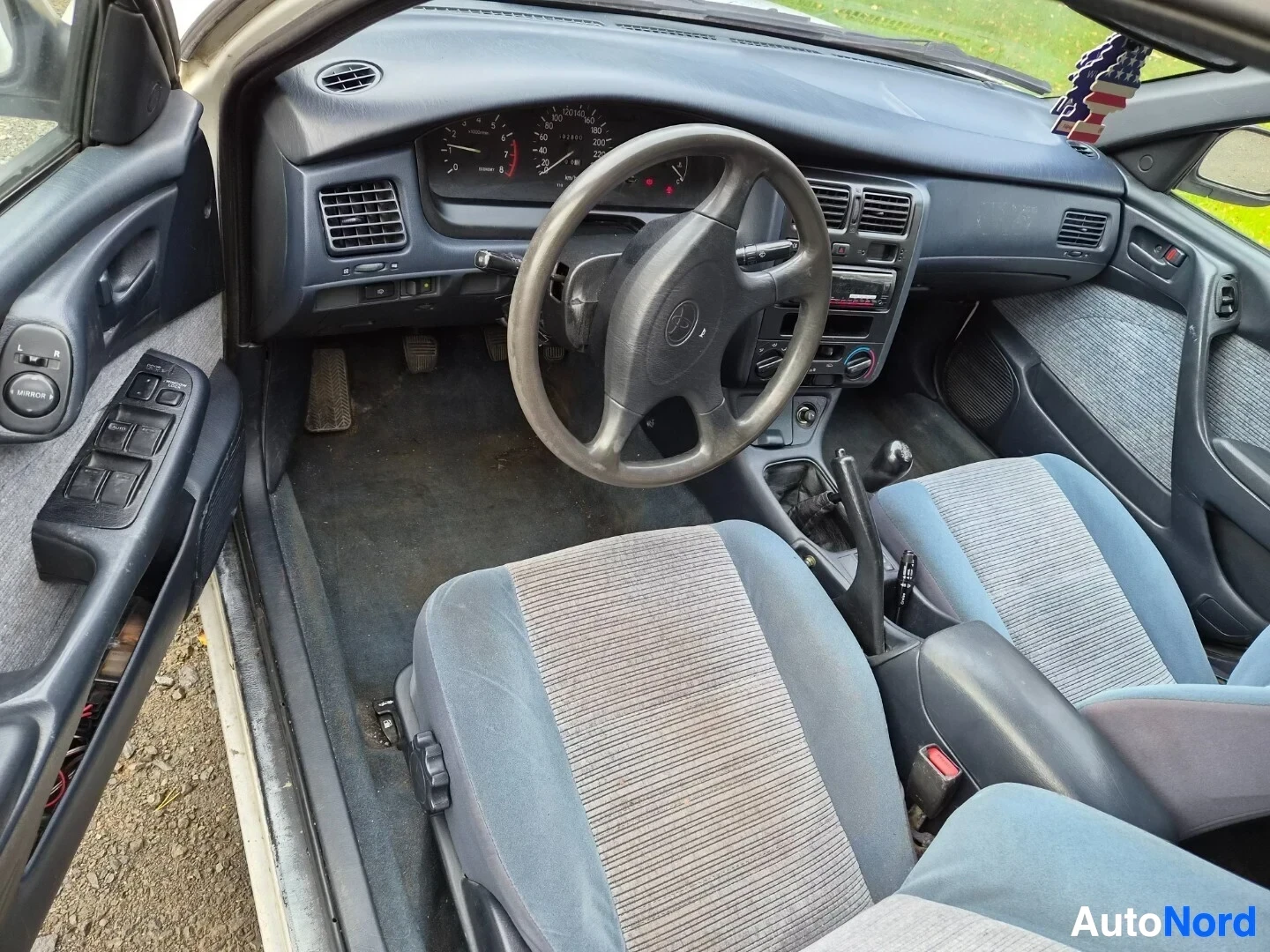 Ohjaamo: Auton ohjaamo kuvattuna kuljettajan puolelta. Näkyvissä on ohjauspyörä, kojelauta, vaihdekeppi ja etuistuimet. Istuimet ovat kangasta, siniset ja harmaaraidalli