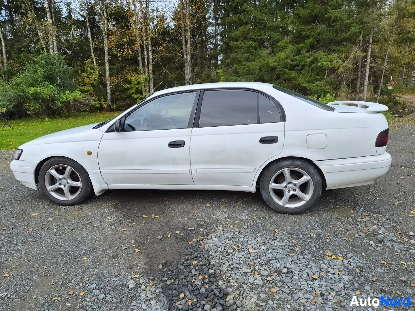Takaviisto: Valkoinen sedan-tyyppinen henkilöauto on pysäköity soratielle vasemmalta sivulta takaviistosta kuvattuna. Autossa on spoileri takaluukussa.
