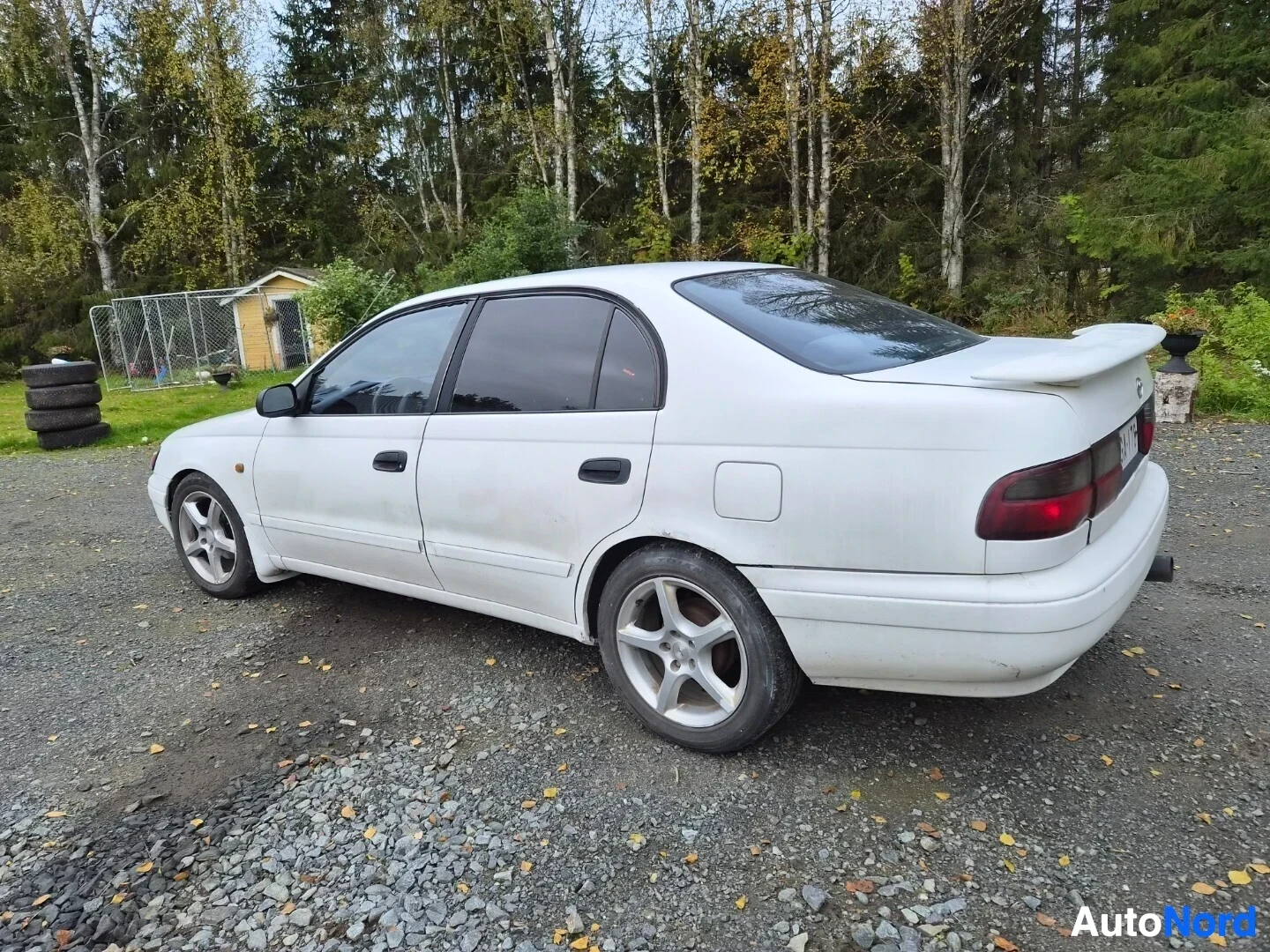 Takaviisto: Valkoinen sedan-tyyppinen auto parkissa sora-alustalla taka-vasemmasta kulmasta kuvattuna. Autossa on spoileri takaluukussa ja tummennetut takaovien ikkunat.