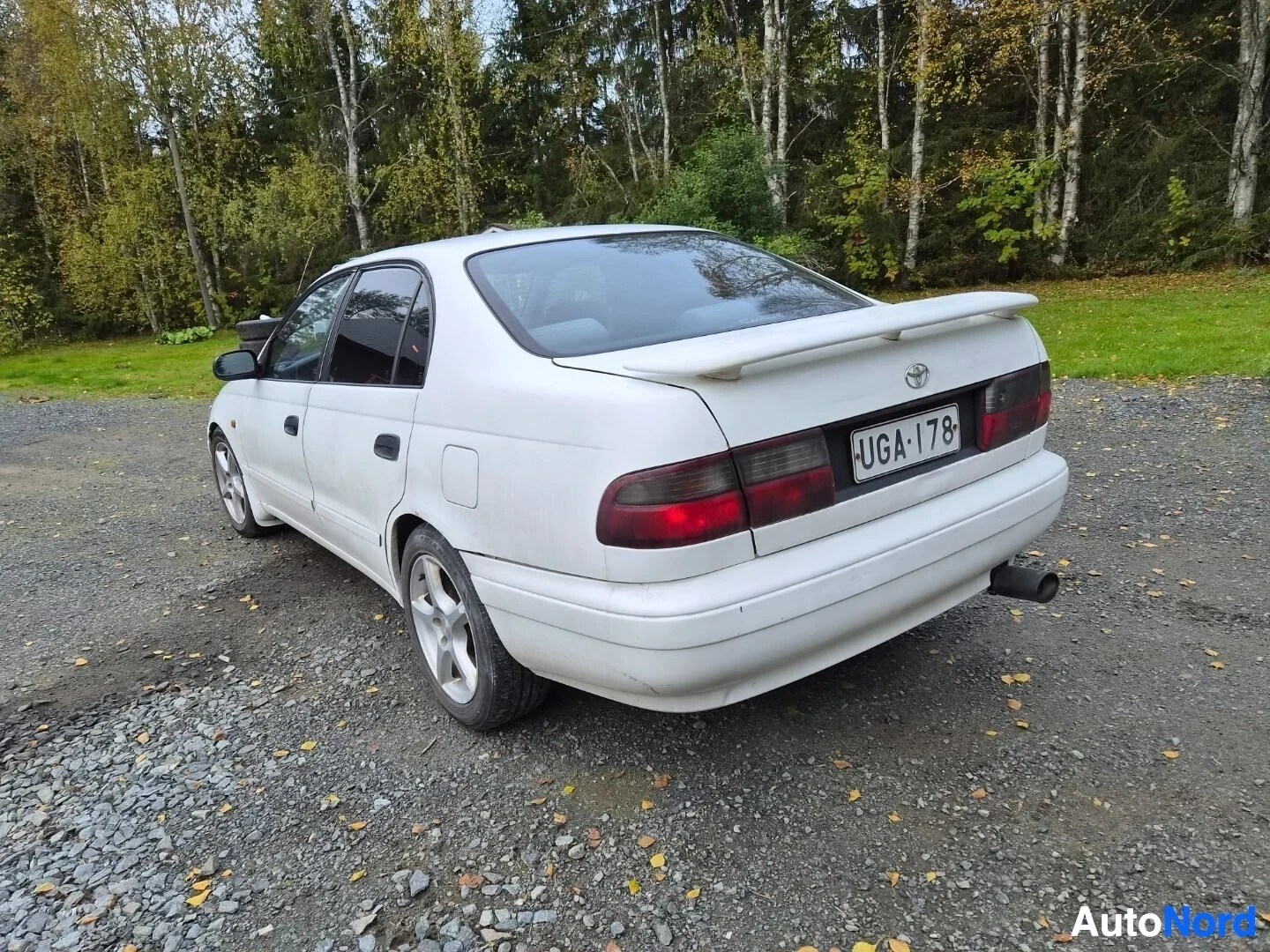 Takaviisto: Valkoinen sedan-tyyppinen auto parkissa sora-alustalla taka-oikeasta kulmasta kuvattuna. Autossa on spoileri takaluukussa, rekisterikilpi "" ja tummennet