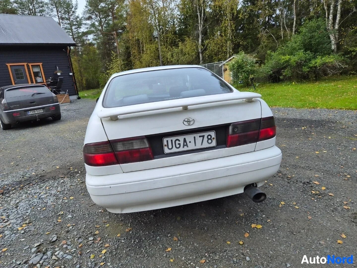 Takaa: Valkoinen sedan-tyyppinen auto parkissa sora-alustalla takaa kuvattuna. Autossa on spoileri takaluukussa, rekisterikilpi "" ja pakoputki näkyy oikealla t