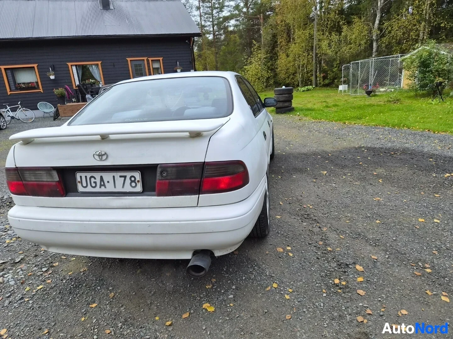 Takaa: Valkoinen sedan-tyyppinen auto parkissa sora-alustalla takaa kuvattuna. Autossa on spoileri takaluukussa, rekisterikilpi "" ja pakoputki näkyy oikealla t