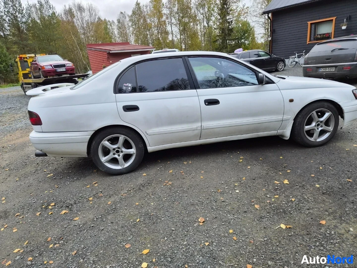 Sivulta: Valkoinen sedan näkyy sivusta, pysäköitynä sorapihalle. Autossa on tummennetut ikkunat ja metallivanteet. Taustalla näkyy punainen pakettiauto hinausauton laval