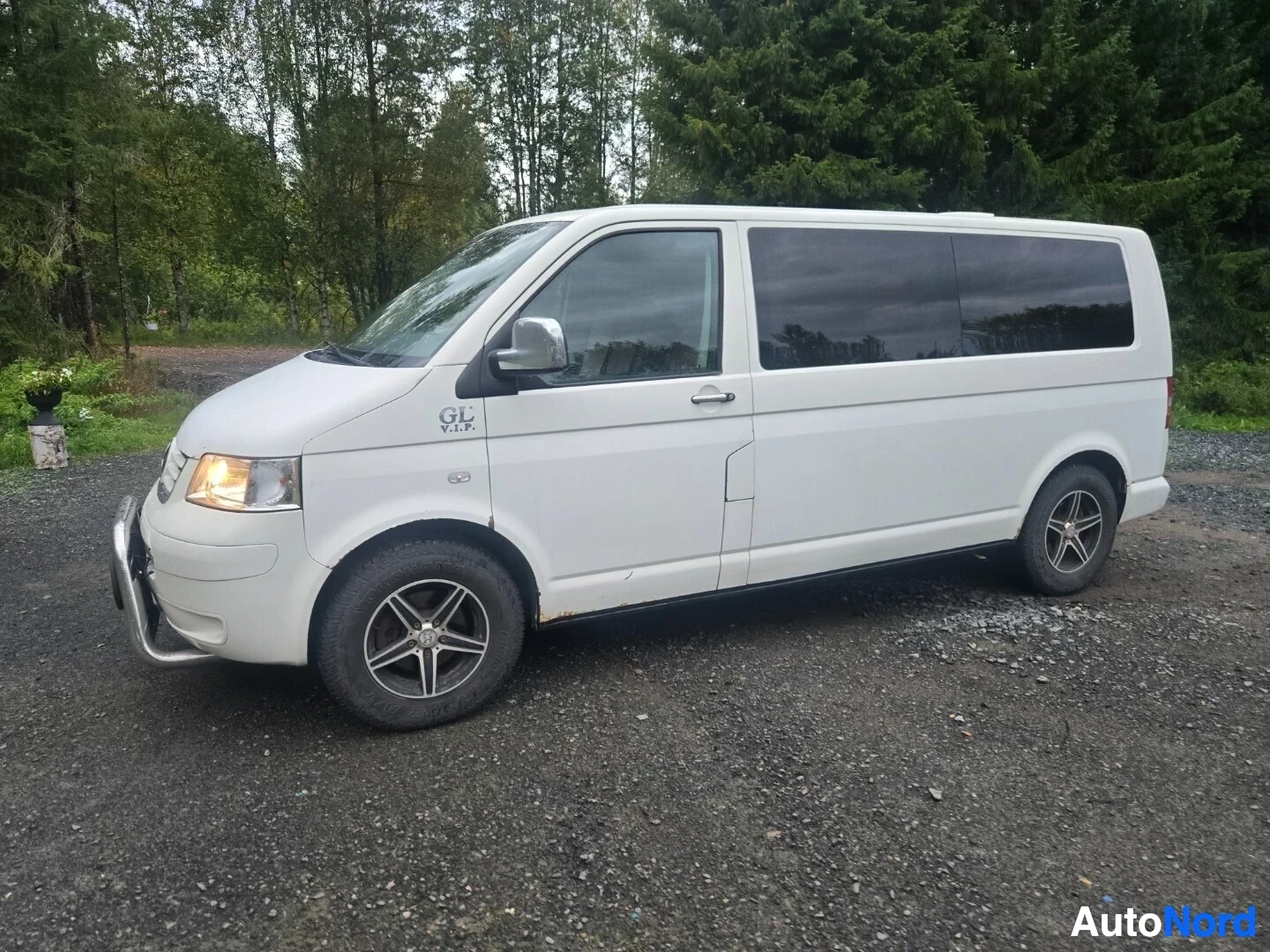 Etuviisto, nokka oikealle: Valkoinen tila-auto, jossa on etupuskurissa metallinen karjarauta, on pysäköity sora-alustalle etuoikealta kuvattuna. Autossa on hopeanväriset vanteet ja