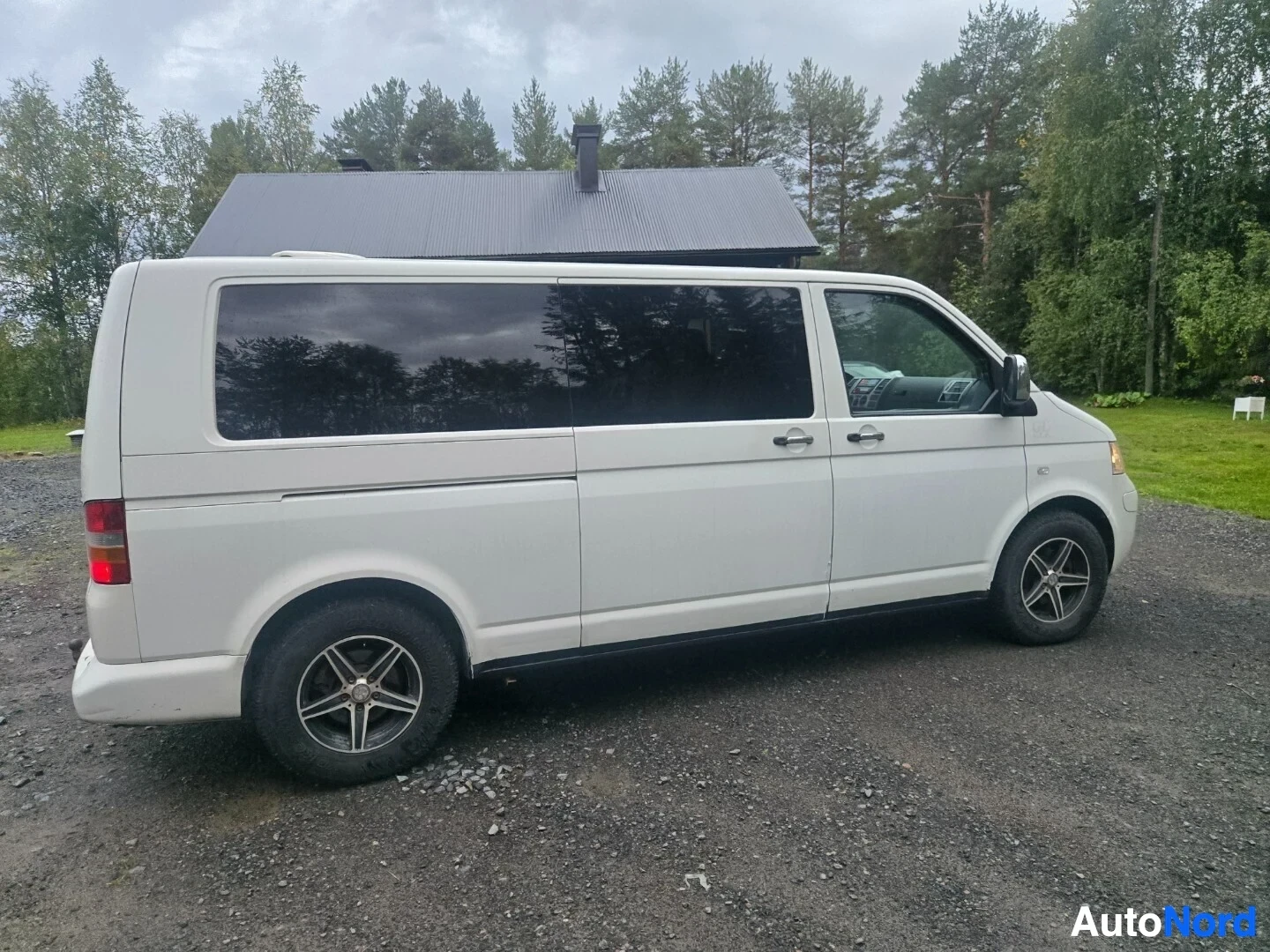 Sivulta: Valkoinen pakettiauto, jossa on mustat ikkunat, on pysäköity soratielle. Kuvassa näkyy auton oikea kylki.