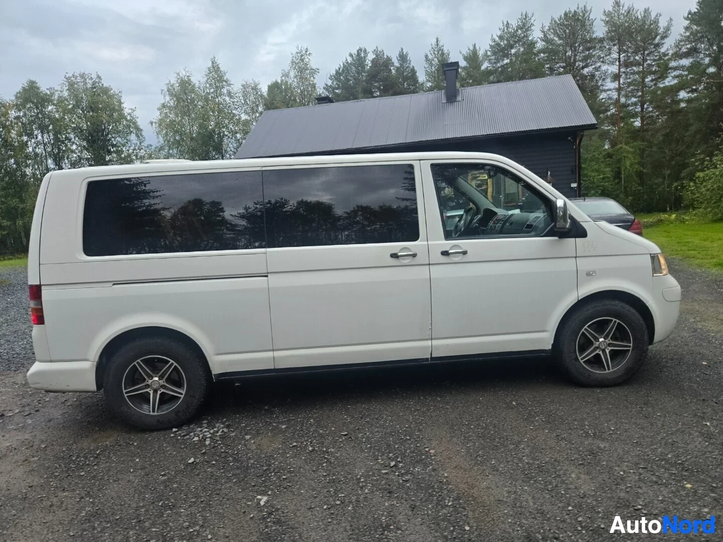 Sivulta: Valkoinen pakettiauto, jossa on mustat ikkunat, on pysäköity soratielle. Kuvassa näkyy auton oikea kylki.