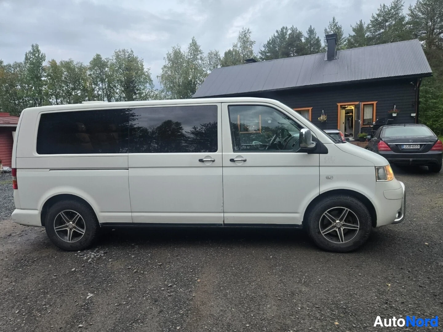 Sivulta: Valkoinen tila-auto pysäköitynä sivuttain sorapihalle talon eteen. Autossa on tummennetut lasit ja alumiinivanteet.