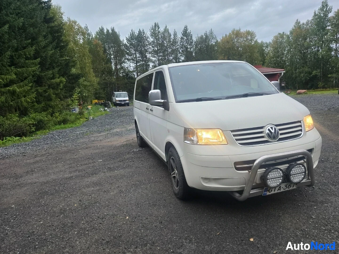 Etuviisto, nokka vasemmalle: Valkoinen tila-auto pysäköitynä keula kohti kuvaajaa sorapihalla. Auto on kallellaan vasemmalle ja sen etuvaloissa on oranssinvärinen heijastus. Auton