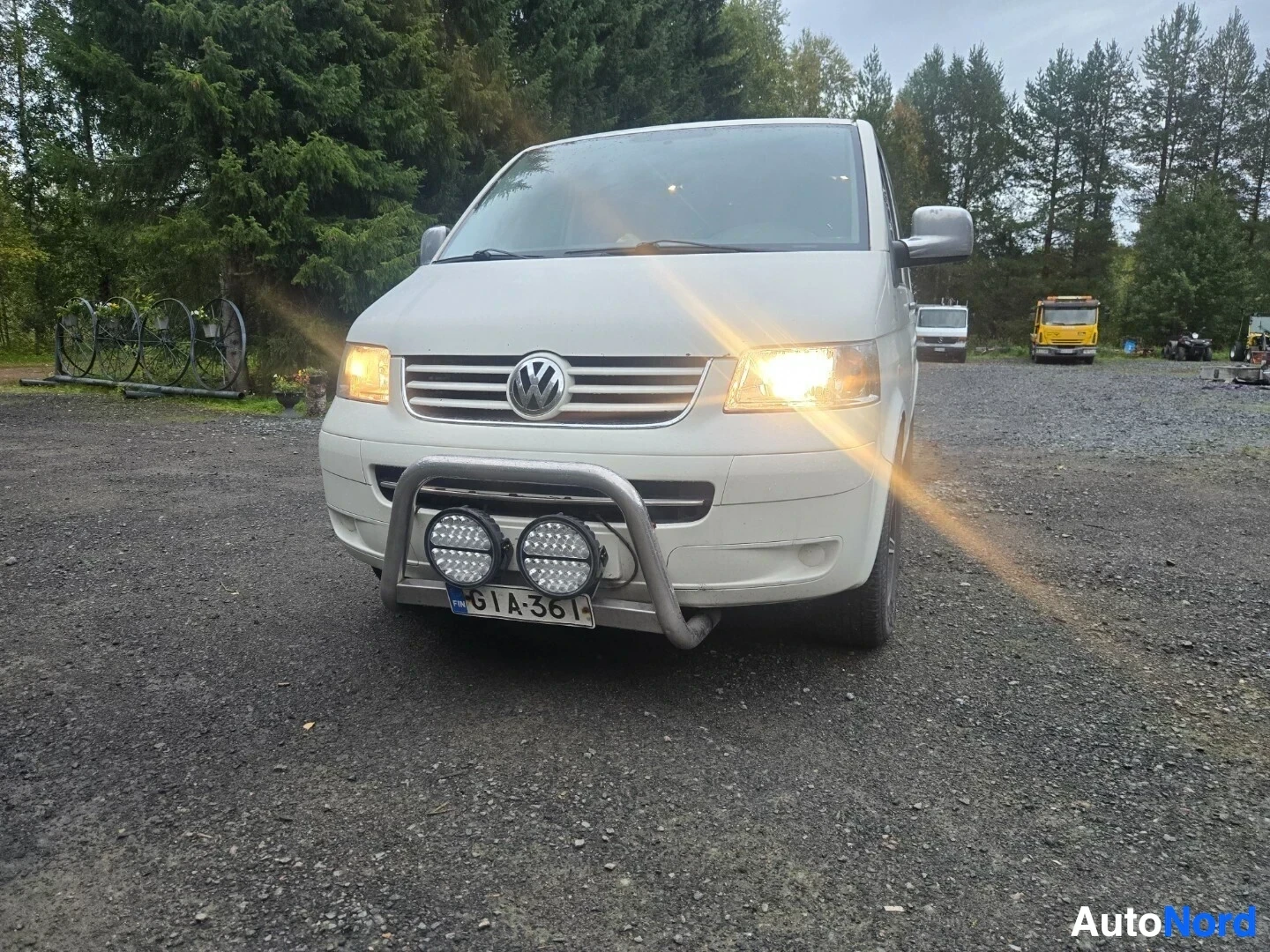 Edestä: Vaalea pakettiauto edestäpäin kuvattuna, ajovalot päällä. Auton keulalla on metallinen karjarauta, johon on kiinnitetty kaksi suurta pyöreää lisävaloa. Rekister