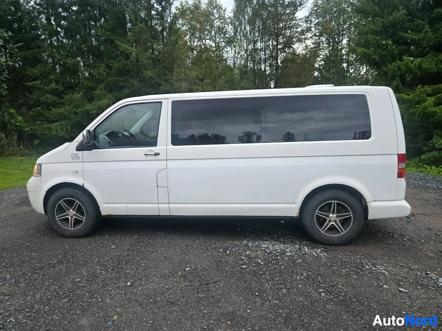 Sivulta: Valkoinen tila-auto, jossa on etupuskurissa metallinen karjarauta, on pysäköity sora-alustalle oikealta sivulta kuvattuna. Autossa on hopeanväriset vanteet ja "