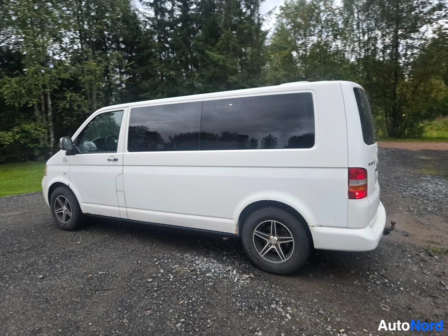 Takaviisto: Valkoinen tila-auto kuvattuna sorapihalla, jossa on tummennetut sivuikkunat. Auton perässä näkyy vetokoukku.
