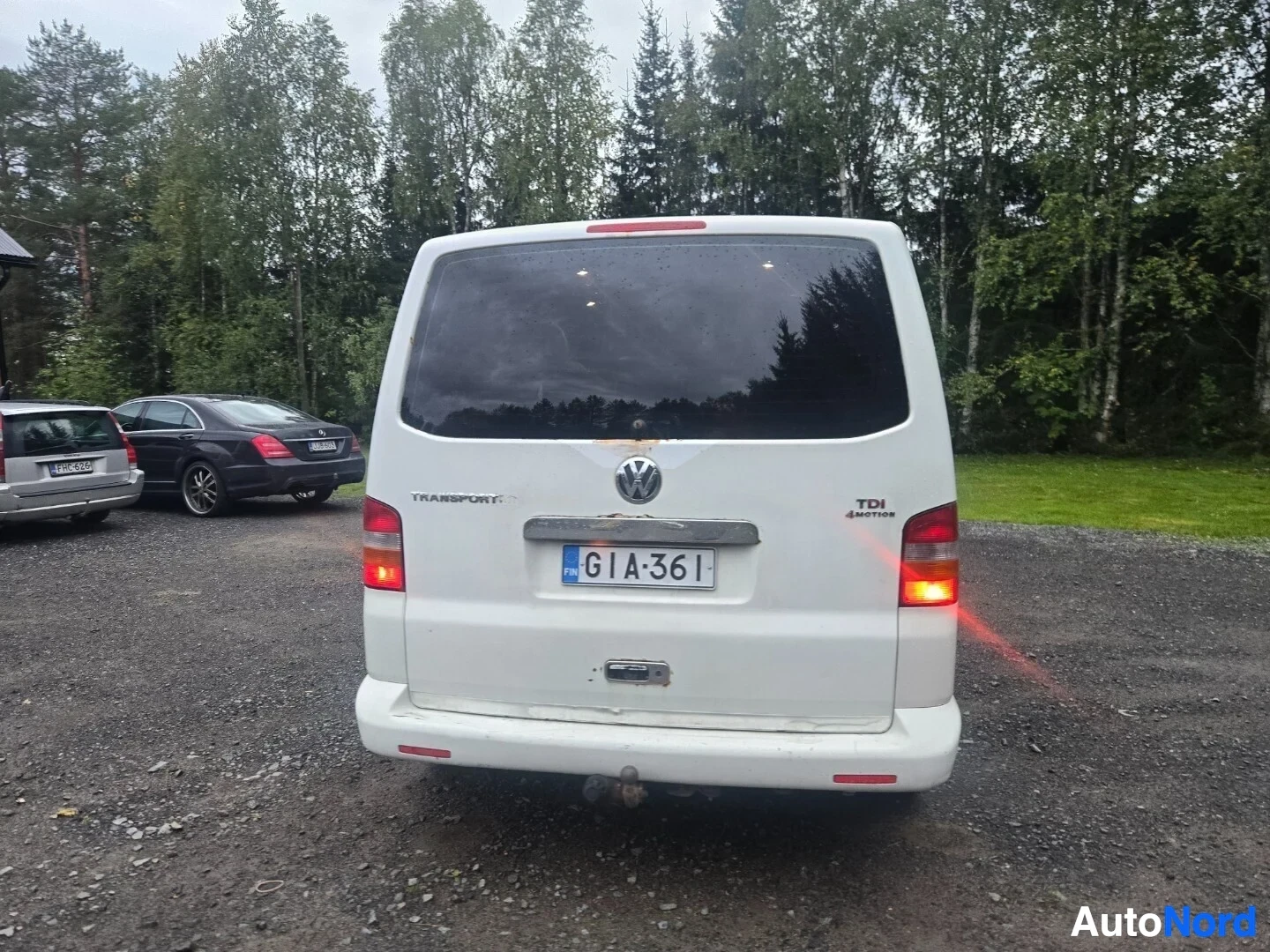Takaa: Valkoinen tila-auto kuvattuna takaa. Takaluukussa on Volkswagen-logo, 'TRANSPORTER' teksti, 'TDI' ja '4MOTION' merkinnät. Rekisterikilpi on keskellä
