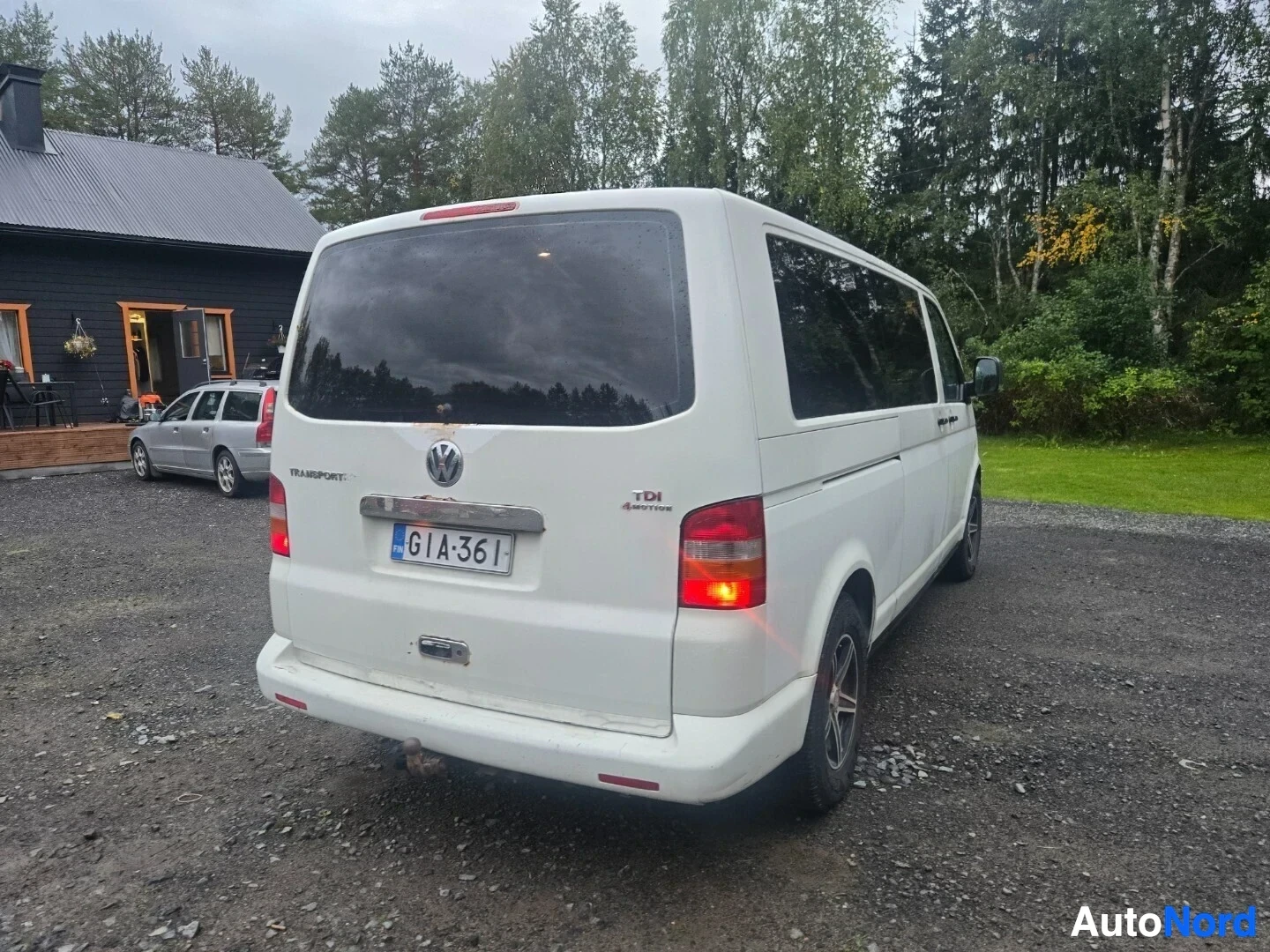 Takaviisto: Valkoinen pakettiauto, jossa on rekisterikilpi ja vetokoukku, on pysäköity soratielle puisen talon eteen. Kuvassa näkyy auton takaosa ja oikea kylki. Ta