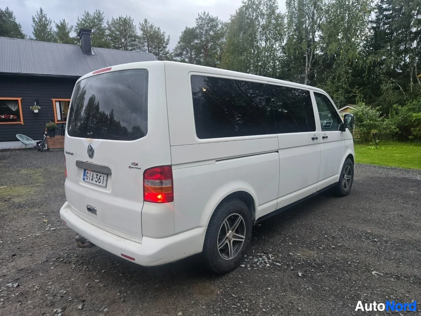 Takaviisto: Valkoinen pakettiauto, jossa on rekisterikilpi ja vetokoukku, on pysäköity soratielle puisen talon eteen. Kuvassa näkyy auton takaosa ja oikea kylki. Ta