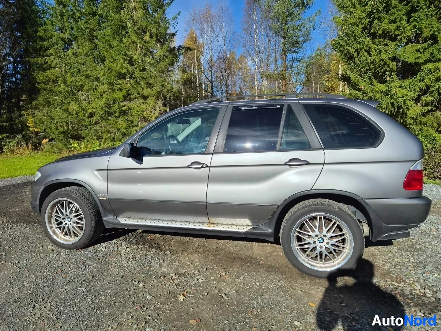 Takaviisto: Harmaa SUV vasemmasta takaviistosta kuvattuna soratiellä. Auton vasen kylki ja takaosa näkyvät. Katolla on kattokiskot ja sivuilla astinlaudat. Autossa on monip